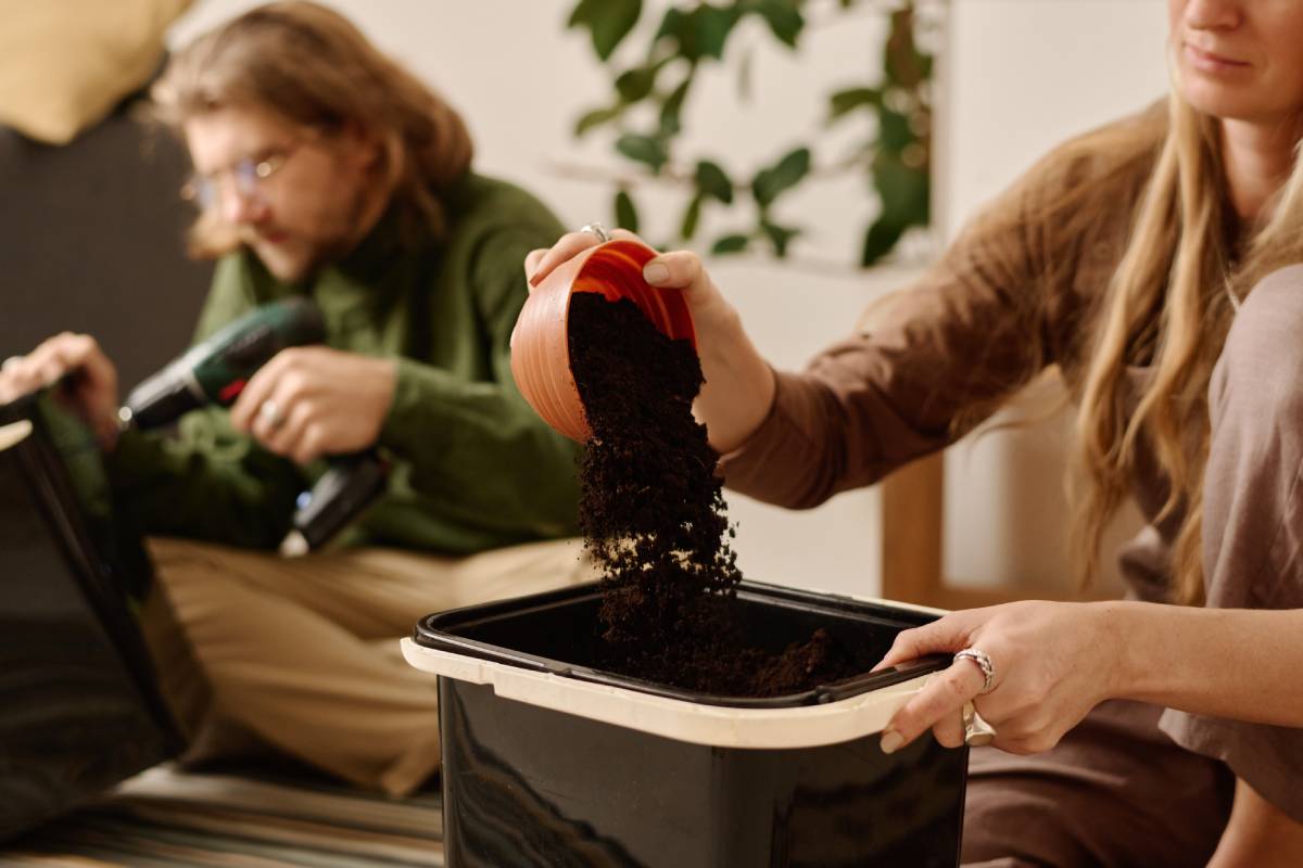 Hands of young woman sprinkling soil from bowl into plastic bucket for food waste against her husband drilling holes in trash bin