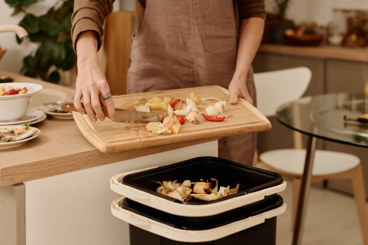 Hands of young housekeeper throwing leftovers of organic meal into trash bin with compost after cooking vegetable salad in the kitchen